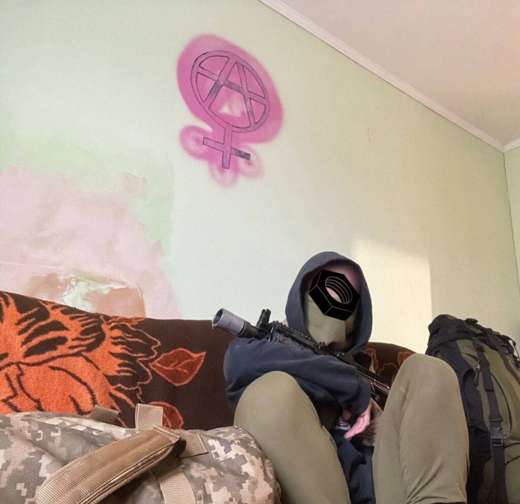 A person dressed in camouflage trousers and a dark hoodie sits on a patterned sofa. They are holding a rifle, and the wall behind them features a pink anarchy symbol combined with the Venus symbol, representing feminism.