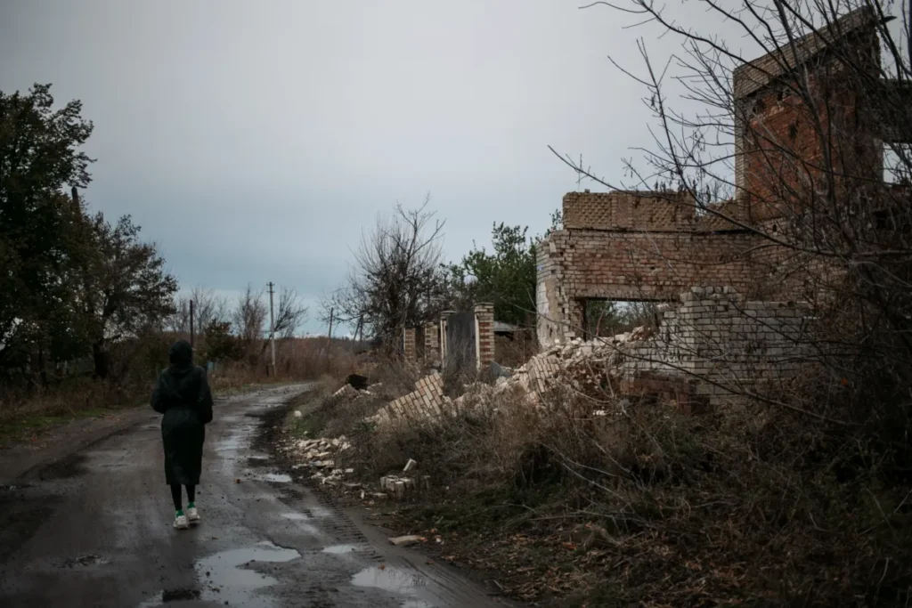 A person walking along a muddy road next to the ruins of a brick building in a rural area, with a cloudy sky overhead.